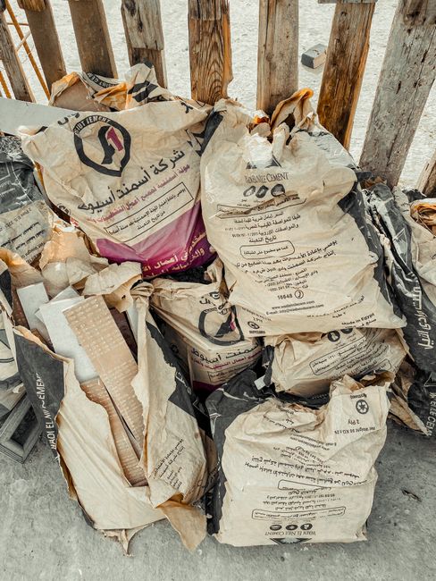 A pile of used cement bags made from thick, woven paper material with printed text and symbols, slightly crumpled and stacked irregularly on an outdoor concrete surface. The bags feature a mix of neut